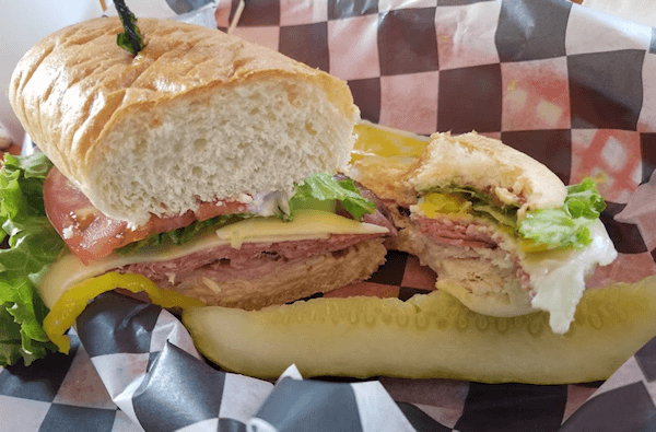 A sliced sub sandwich is displayed on checkered paper, with various fillings and a pickle.