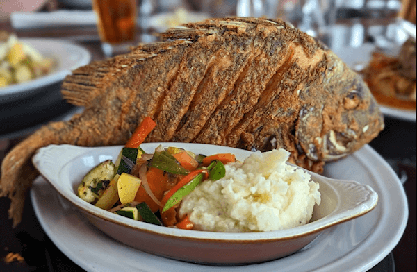 A plate with a whole fried fish with vegetables and mashed potatoes in a side dish 